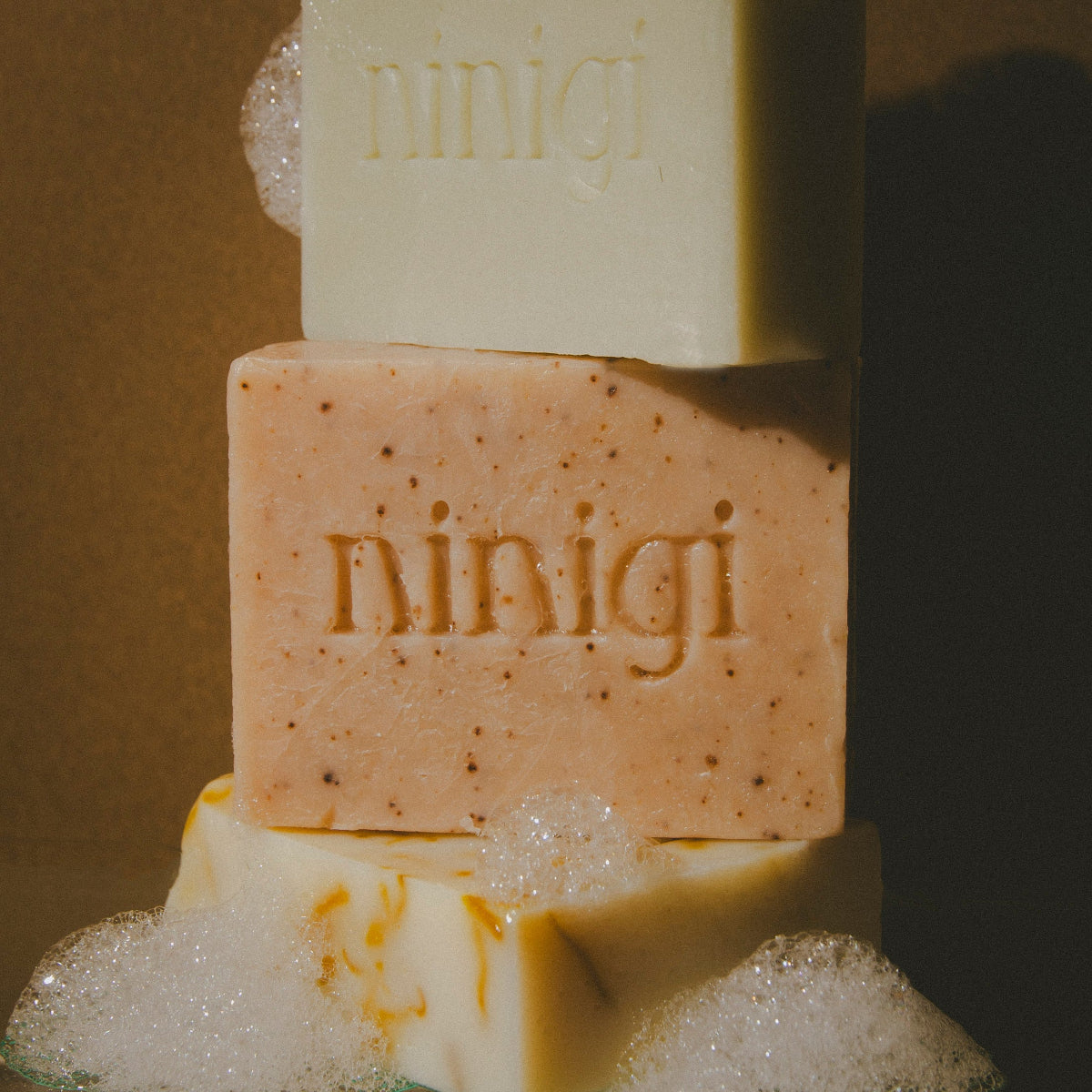 Three bars of soap stacked with 'ninigi' branding, surrounded by bubbles on a dark background.