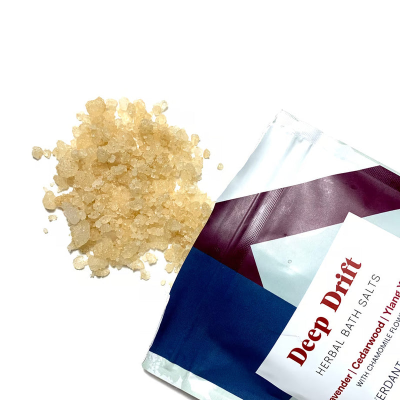A packet of Verdant Alchemy Deep Drift Herbal Bath salts next to a small pile of light beige bath salts. The packet is predominantly white with purple and blue accents and reads 'Deep Drift Herbal Bath Salts'.